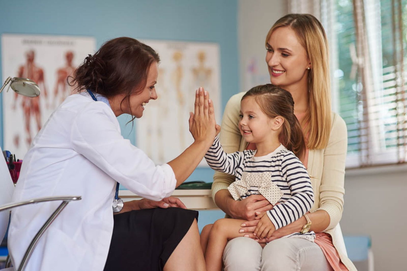 Médico chocando los cinco con una niña durante una cita médica, con la madre presente.