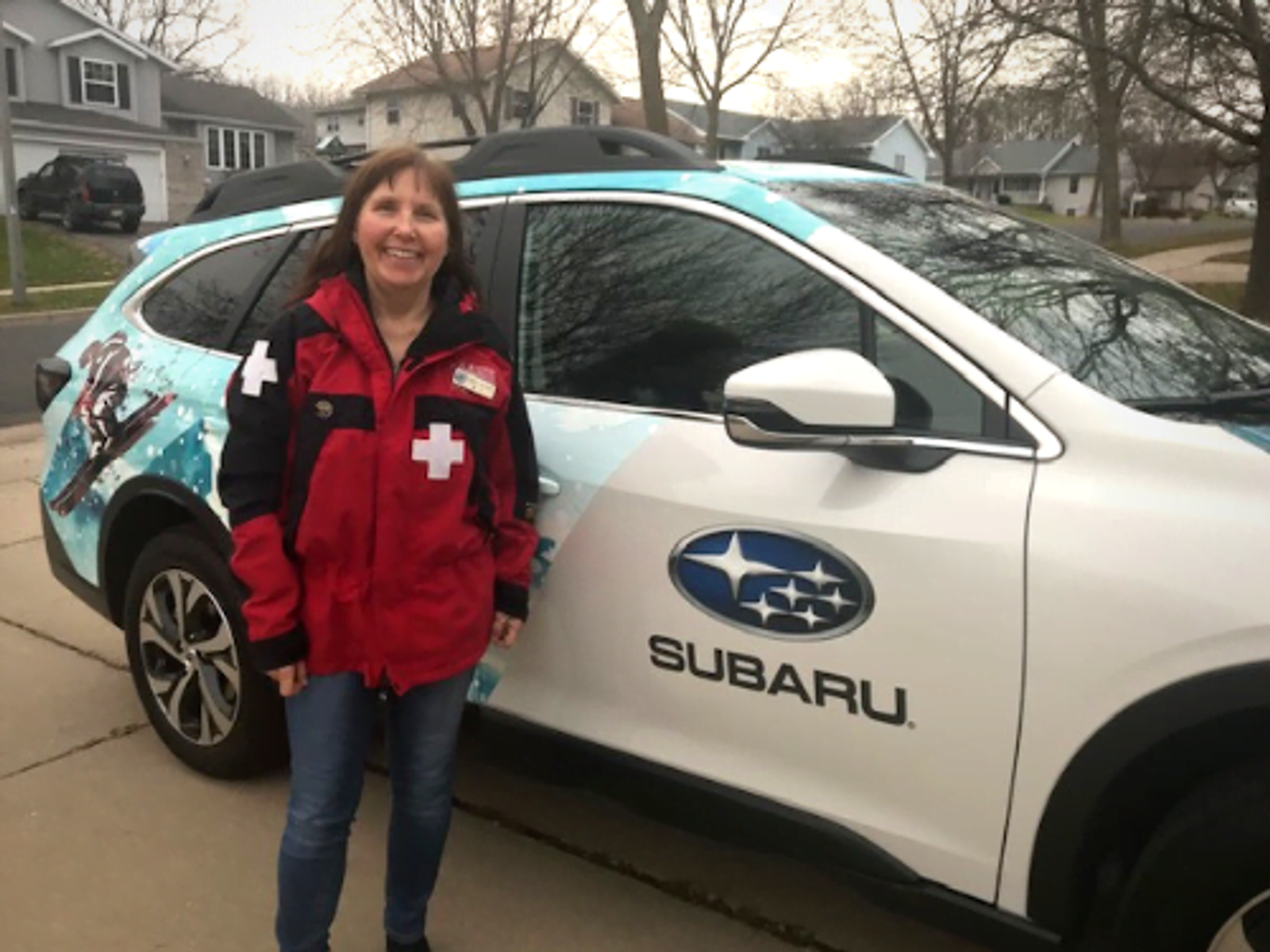 Woman in red jacket standing beside white Subaru with medical cross decal in driveway.