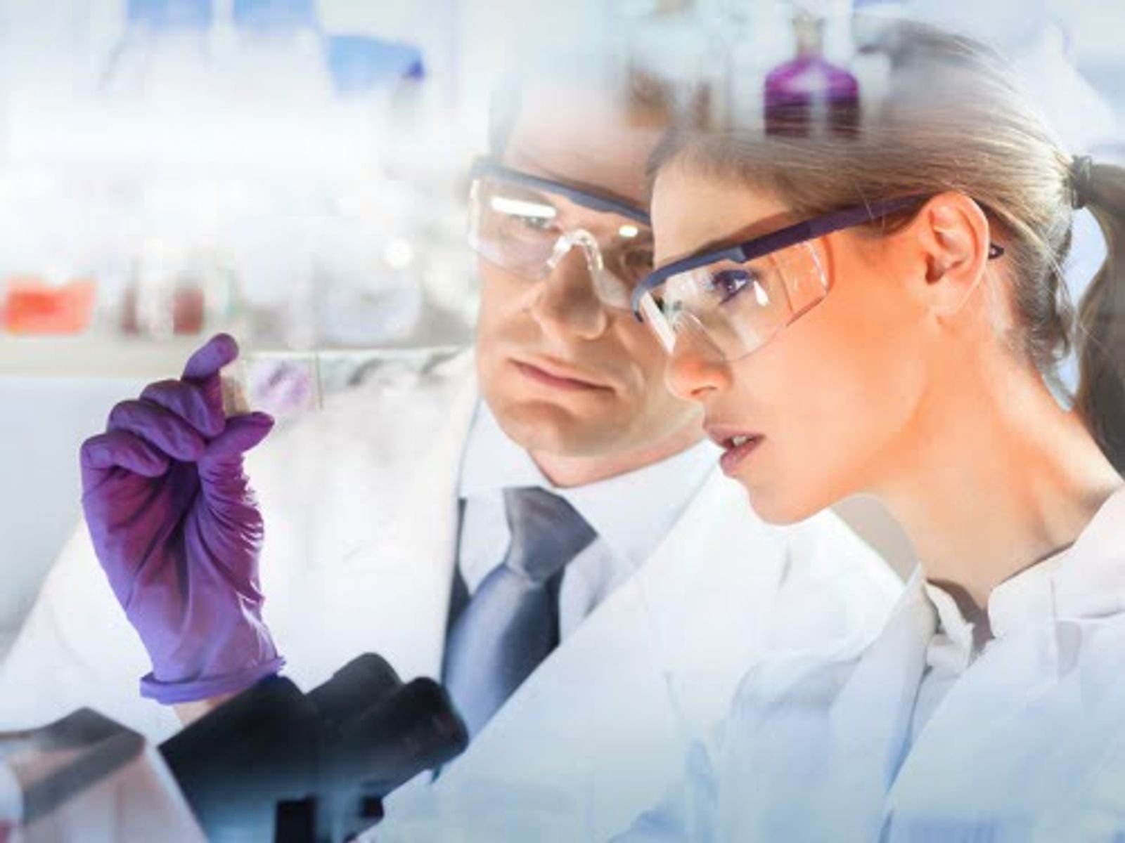 Two scientists in lab coats and safety glasses examining a purple liquid sample in a laboratory setting.