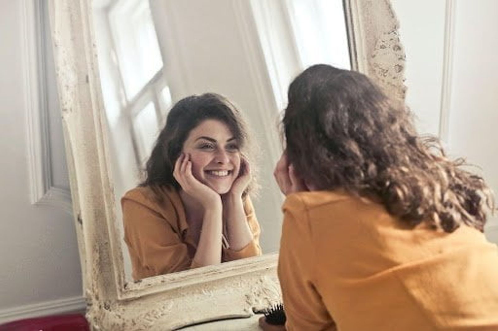 Woman in mustard-yellow shirt smiling at her reflection in an ornate mirror.
