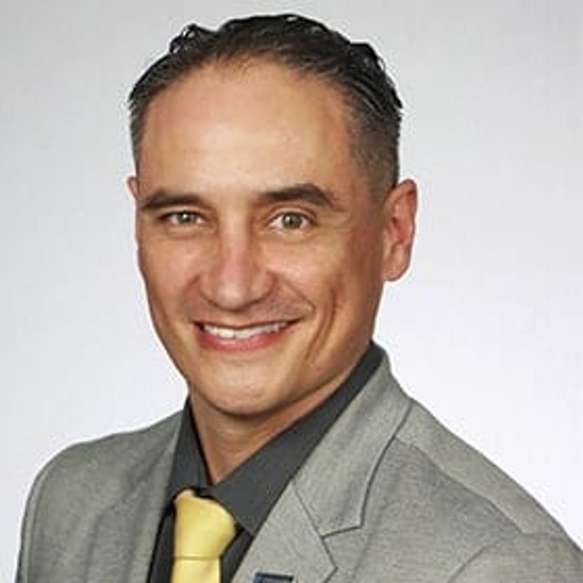 Retrato profesional de un hombre sonriente con saco gris y corbata amarilla, sobre fondo blanco.