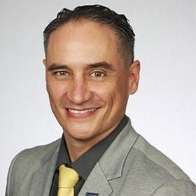 Professional headshot of a smiling man in gray blazer and yellow tie against white background.