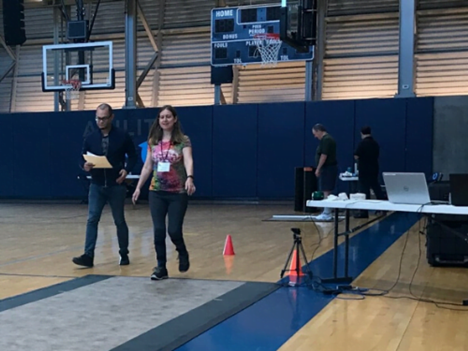 Two people walking on indoor basketball court with equipment and setup tables visible.