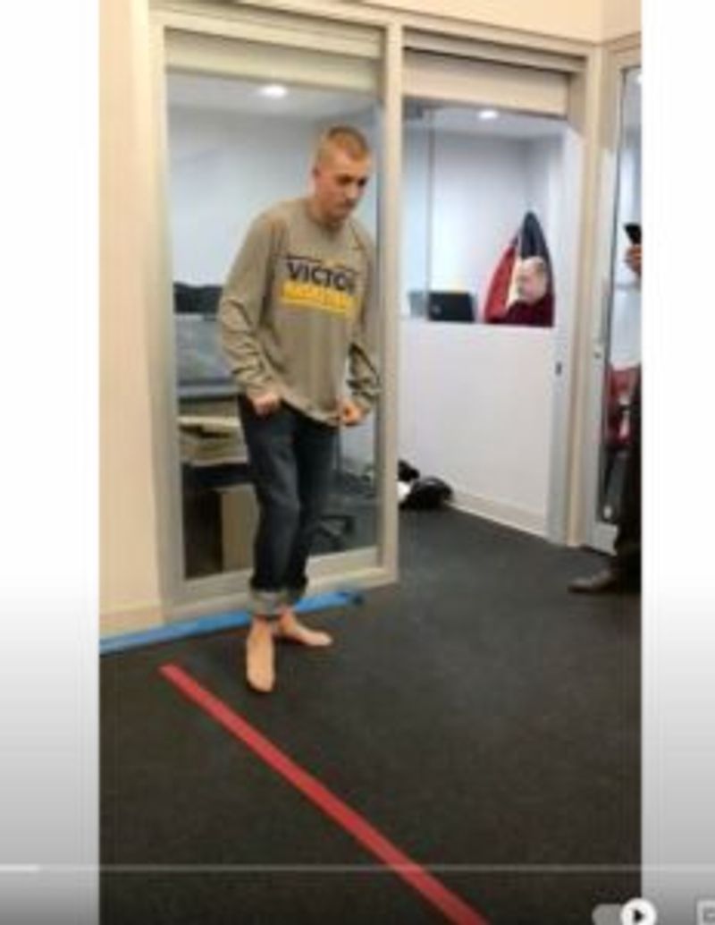 Hombre con sudadera color canela "VICTOR", equilibrándose sobre una pierna cerca de una línea roja en el suelo de un gimnasio.