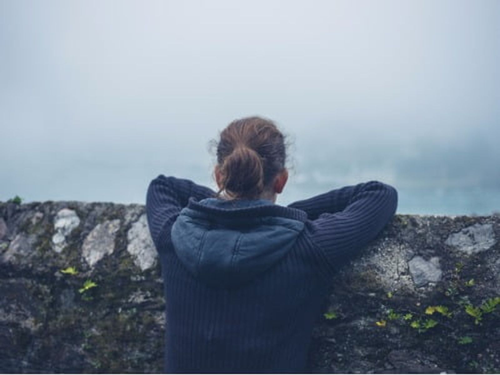 Person with hands behind head sitting on mossy stone wall, facing misty landscape.