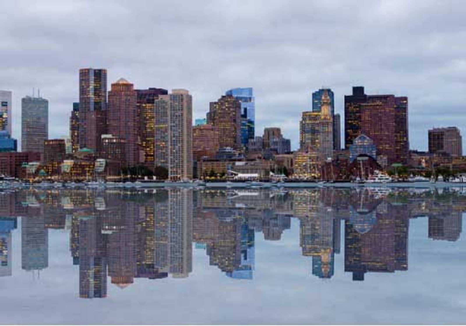 Boston skyline with downtown buildings reflected in calm water at dusk.