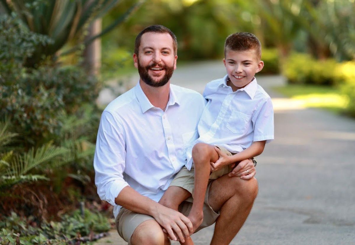 Adult man and young boy smiling together outdoors on a tree-lined path.