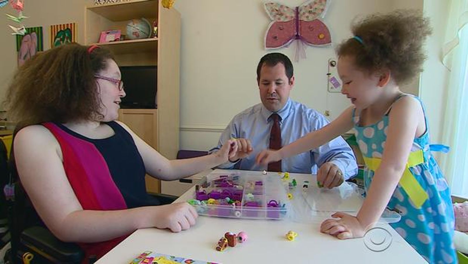 Three people at table with colorful craft supplies and organized storage boxes in background.