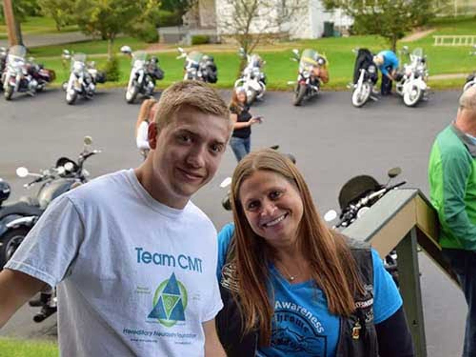 Two smiling people wearing Team CMT shirts stand before parked motorcycles at outdoor event.