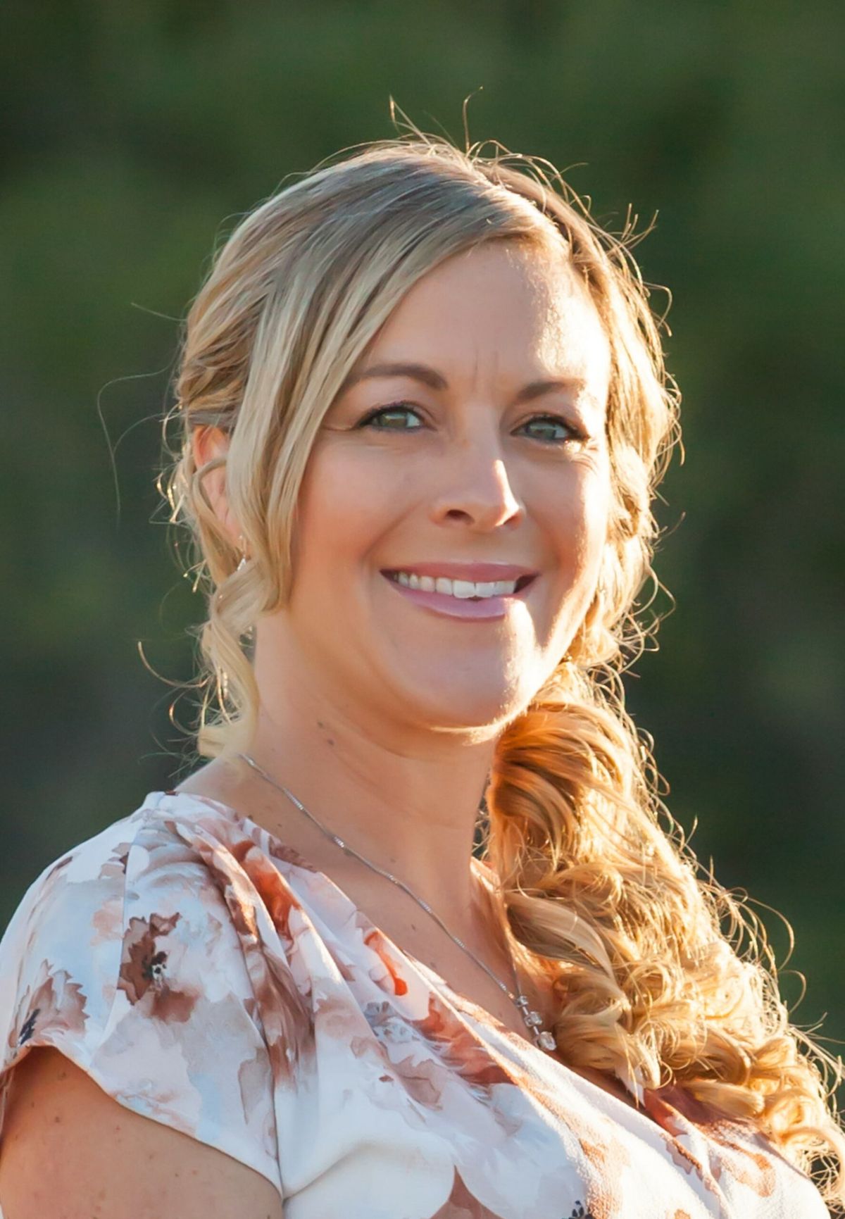 Woman with blonde hair in side braid, smiling at camera, wearing patterned light-colored top, outdoors with blurred green background.