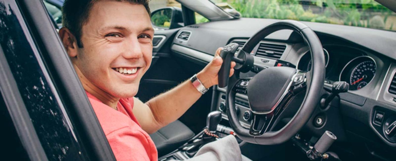 Young man in pink shirt smiling while sitting in driver's seat of car.