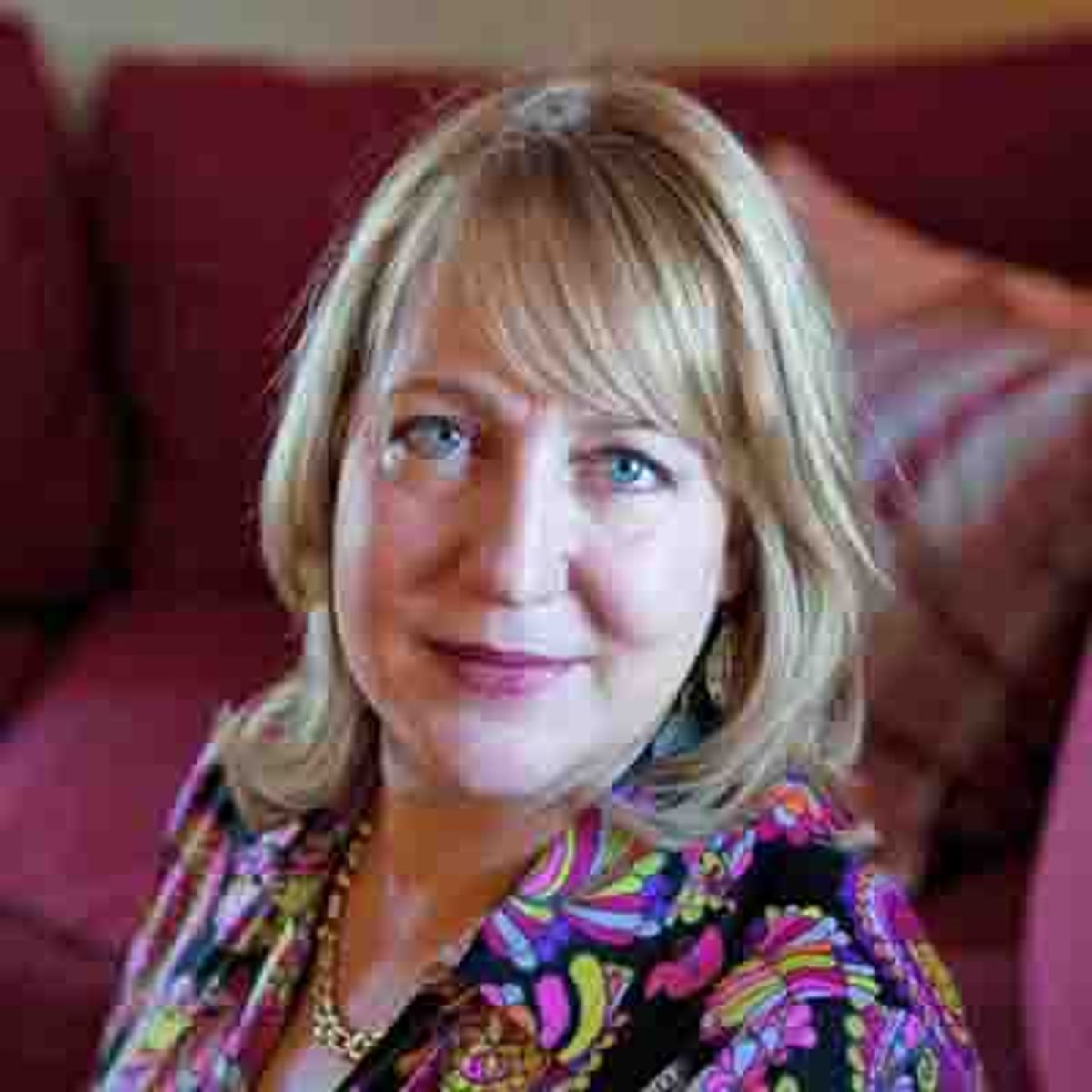 Woman with blonde bangs seated on red couch, wearing colorful patterned blouse.