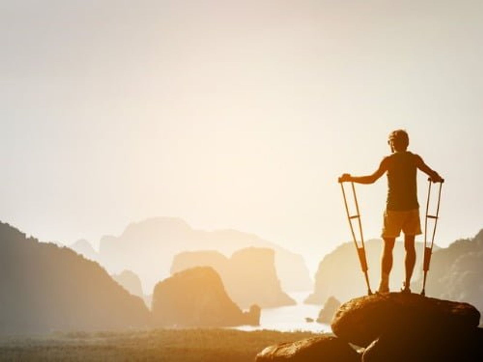 Silhouetted figure with crutches standing on rock overlooking misty mountain landscape at sunrise.
