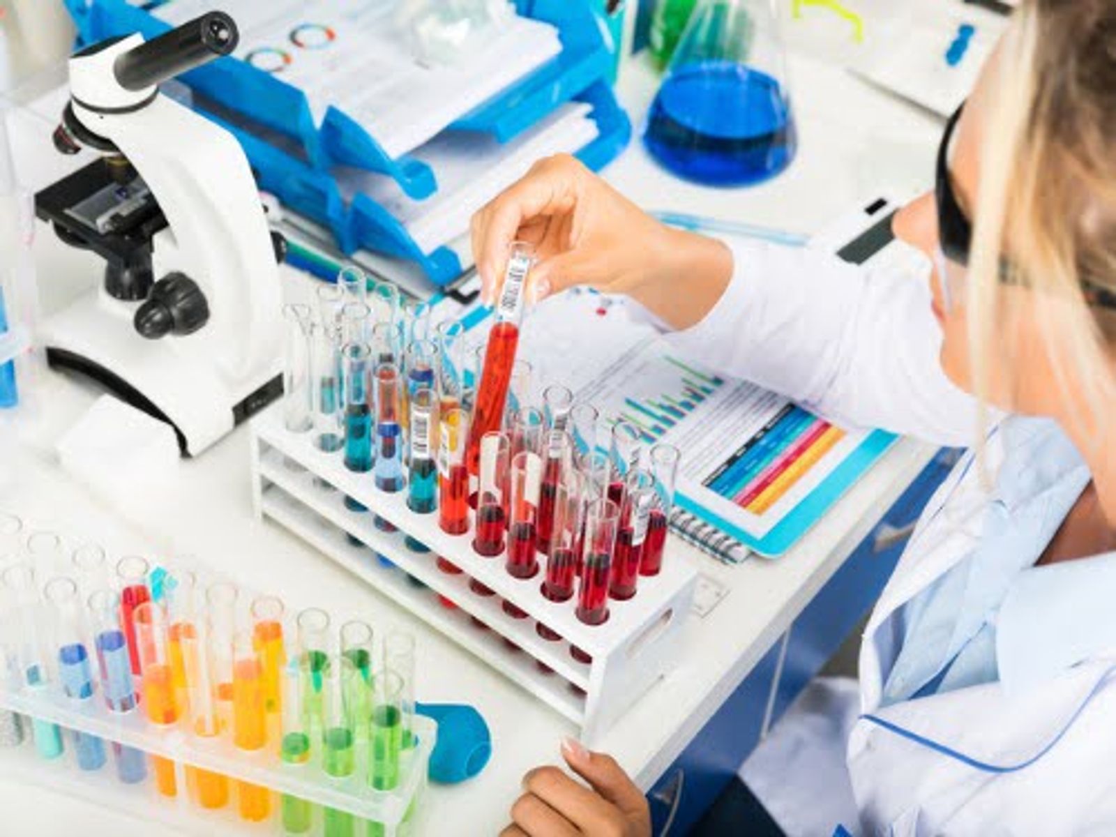 Laboratory technician examining test tubes with colored liquid samples at workstation with microscope.