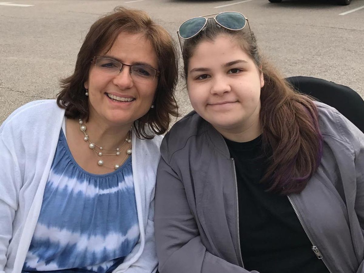 Two smiling women pose together outdoors, one wearing glasses on her head.