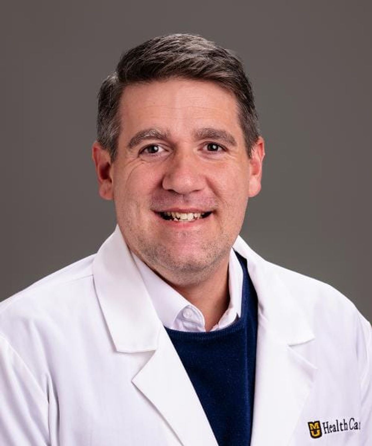 Professional headshot of man in white medical coat with "HealthCare" logo, smiling at camera against gray background.