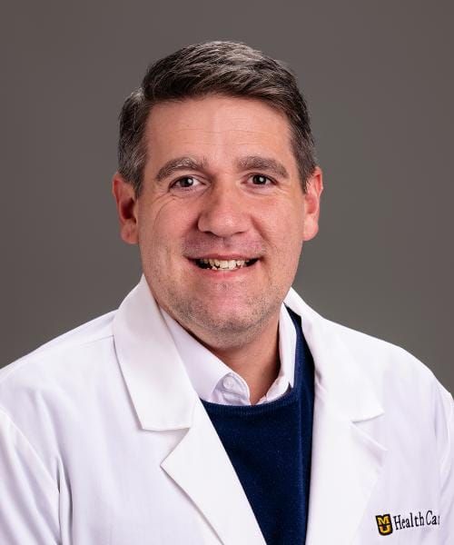 Retrato profesional de un hombre con bata médica blanca con el logotipo "HealthCare", sonriendo a la cámara sobre un fondo gris.