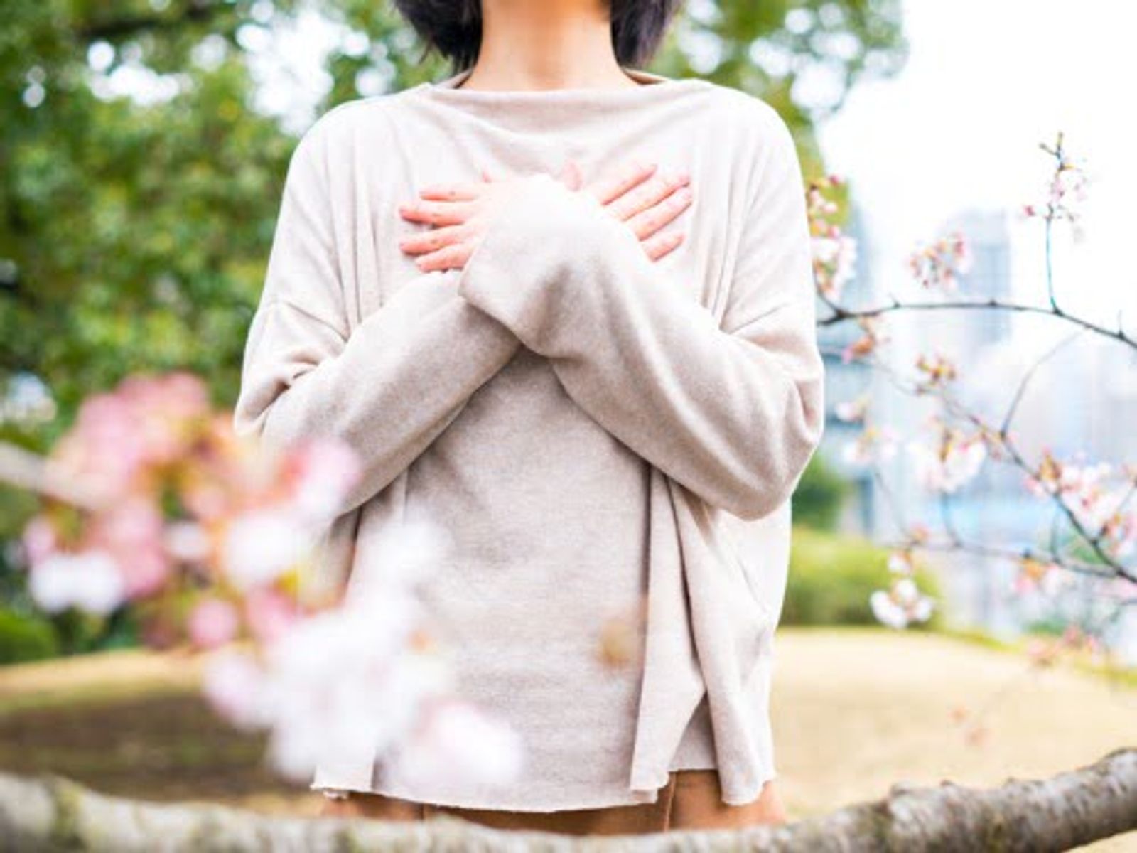 Woman in beige cardigan with arms crossed, standing outdoors among blooming flowers and green foliage.
