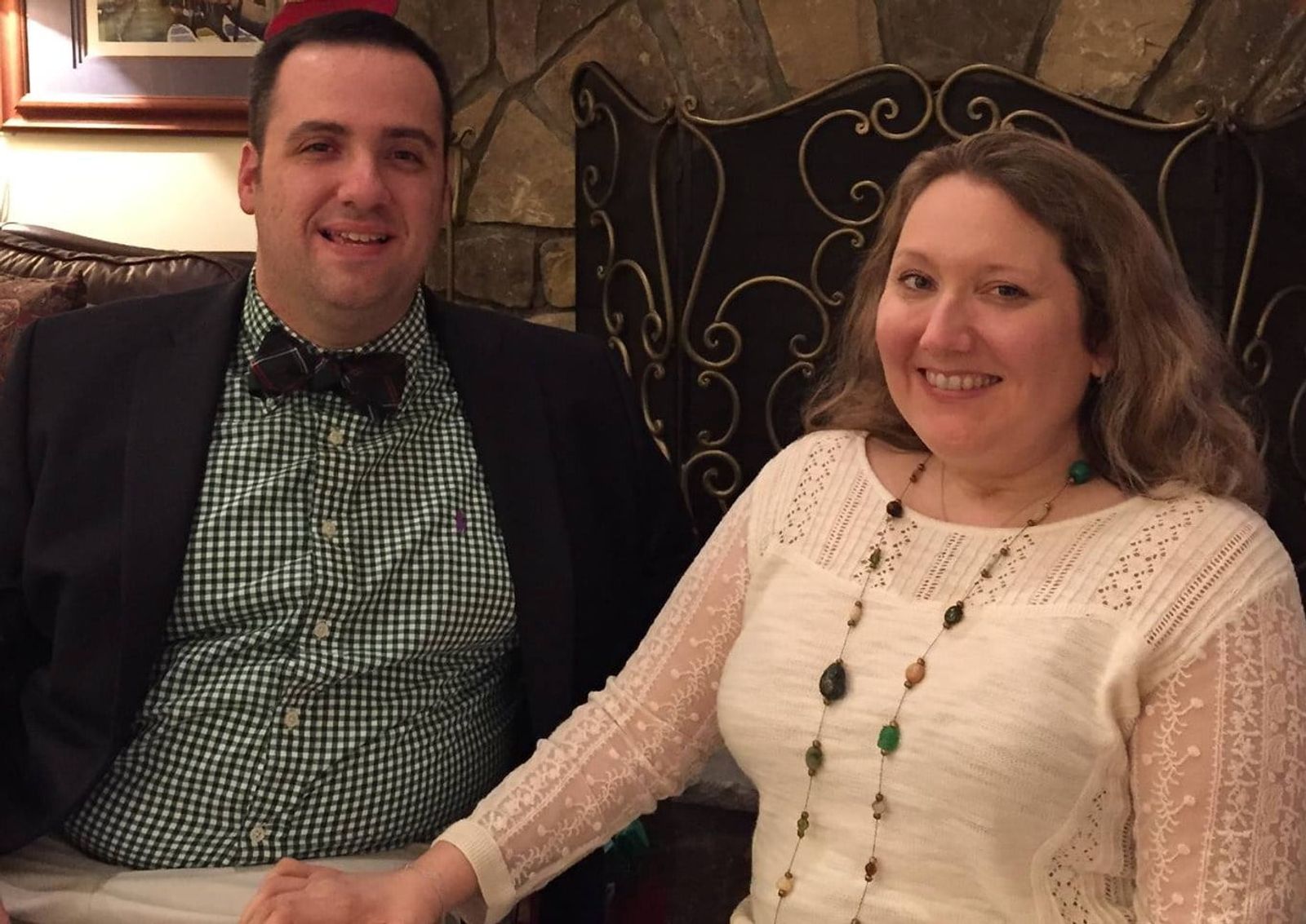 Two people smiling at camera in formal attire indoors with decorative iron chair visible behind them.
