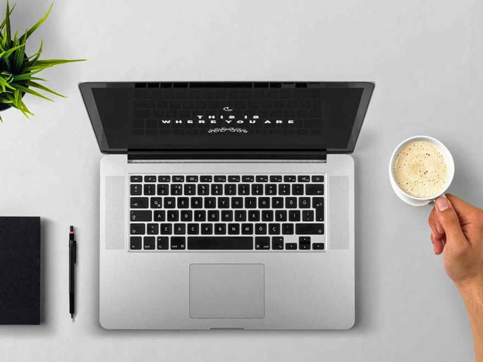 Overhead view of laptop with coffee cup, plant, notebook, and pen on white desk.
