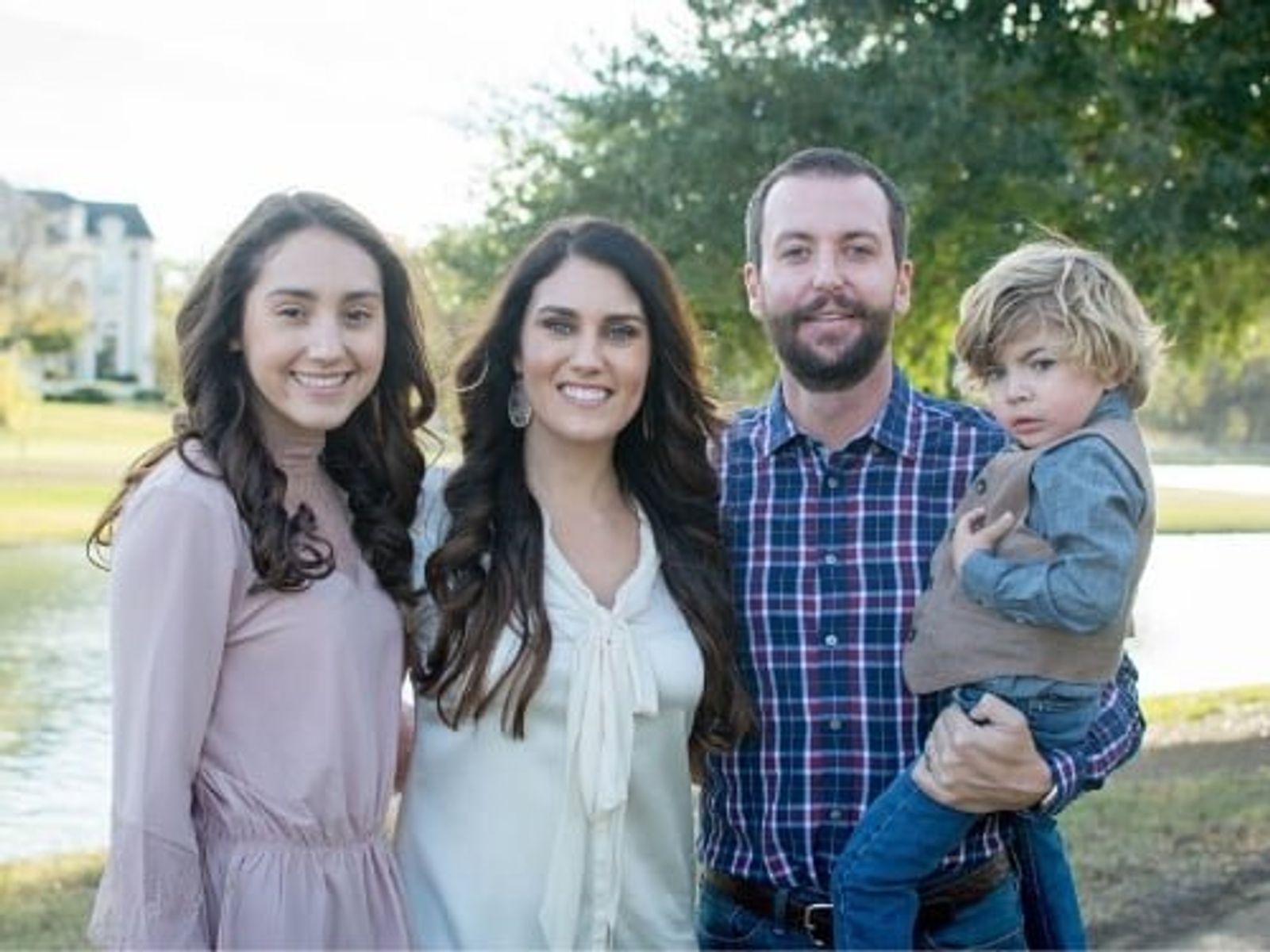 Family of four posing outdoors by a pond with trees and house in background.