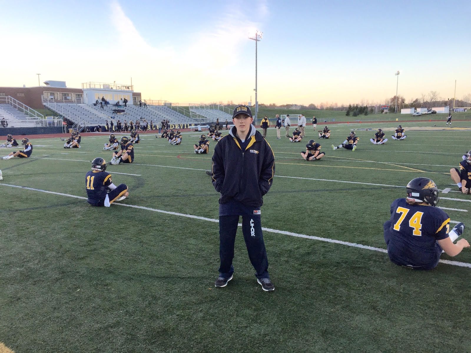 Football coach standing on field with seated players wearing navy uniforms during practice.