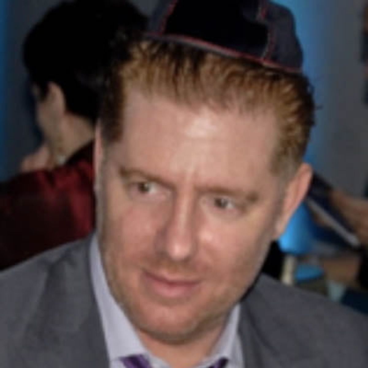 Hombre con traje y corbata morada, con gorra de béisbol roja y negra, en interiores.