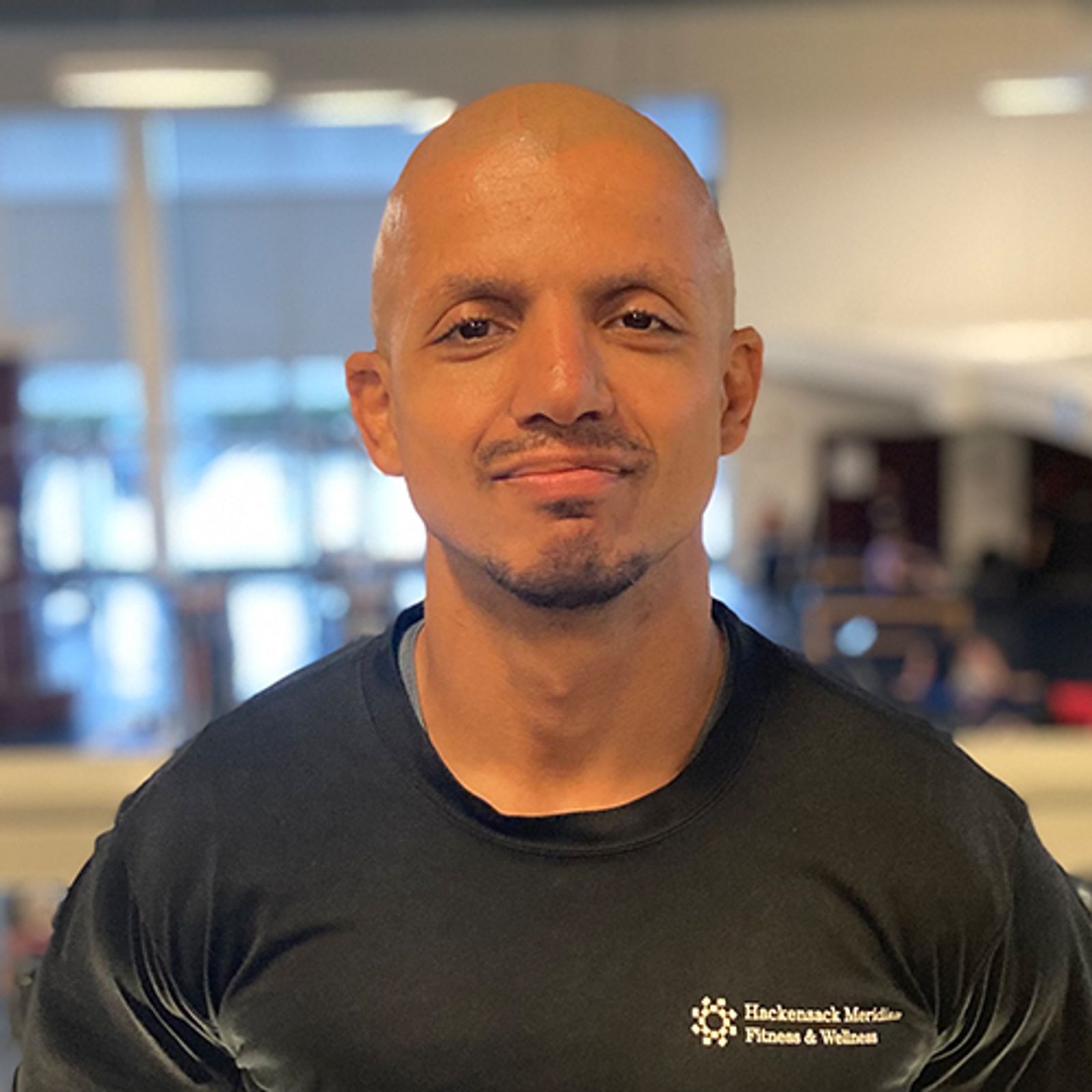 Bald man wearing black t-shirt with "Hackensack Meridian Fitness & Wellness" logo, indoors with windows behind.