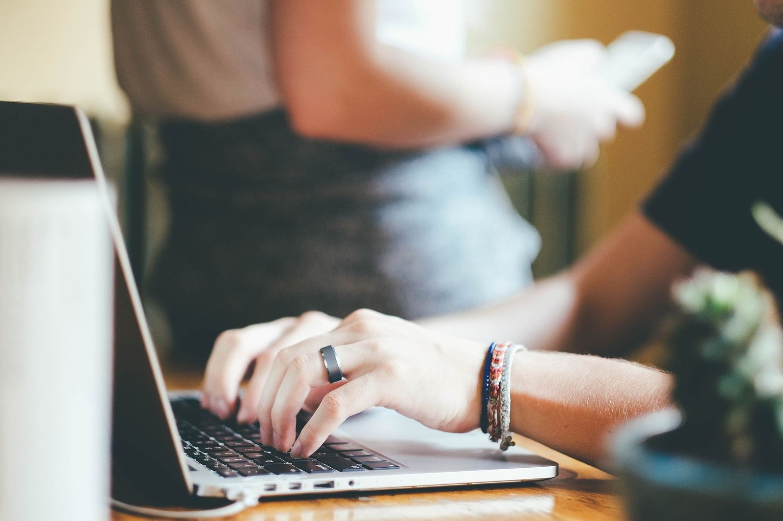 Woman typing on laptop keyboard while wearing rings and bracelets at wooden desk.