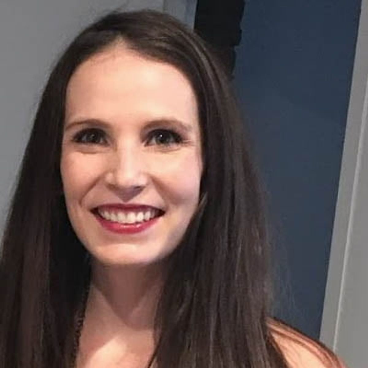 Woman with long dark hair smiling at camera, wearing pink lipstick and orange top.