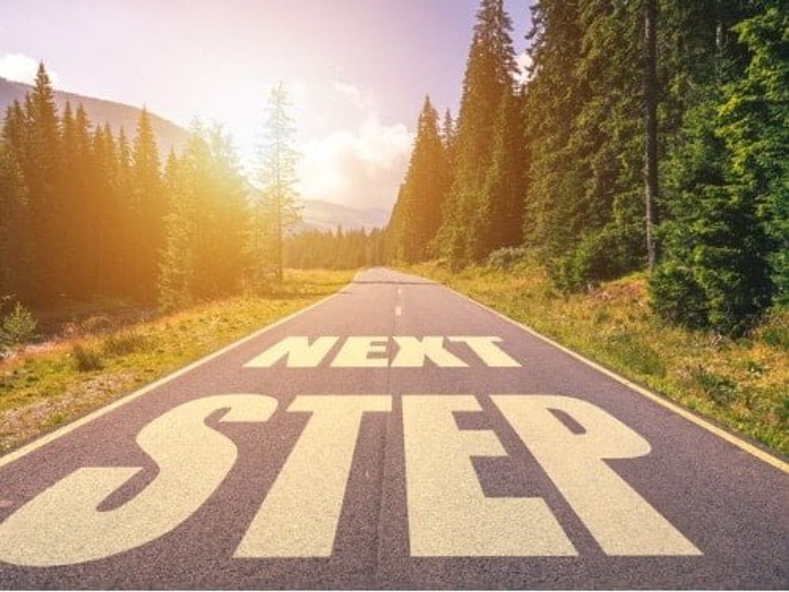 Straight road through forest with "NEXT STEP" painted on asphalt, sunset lighting.