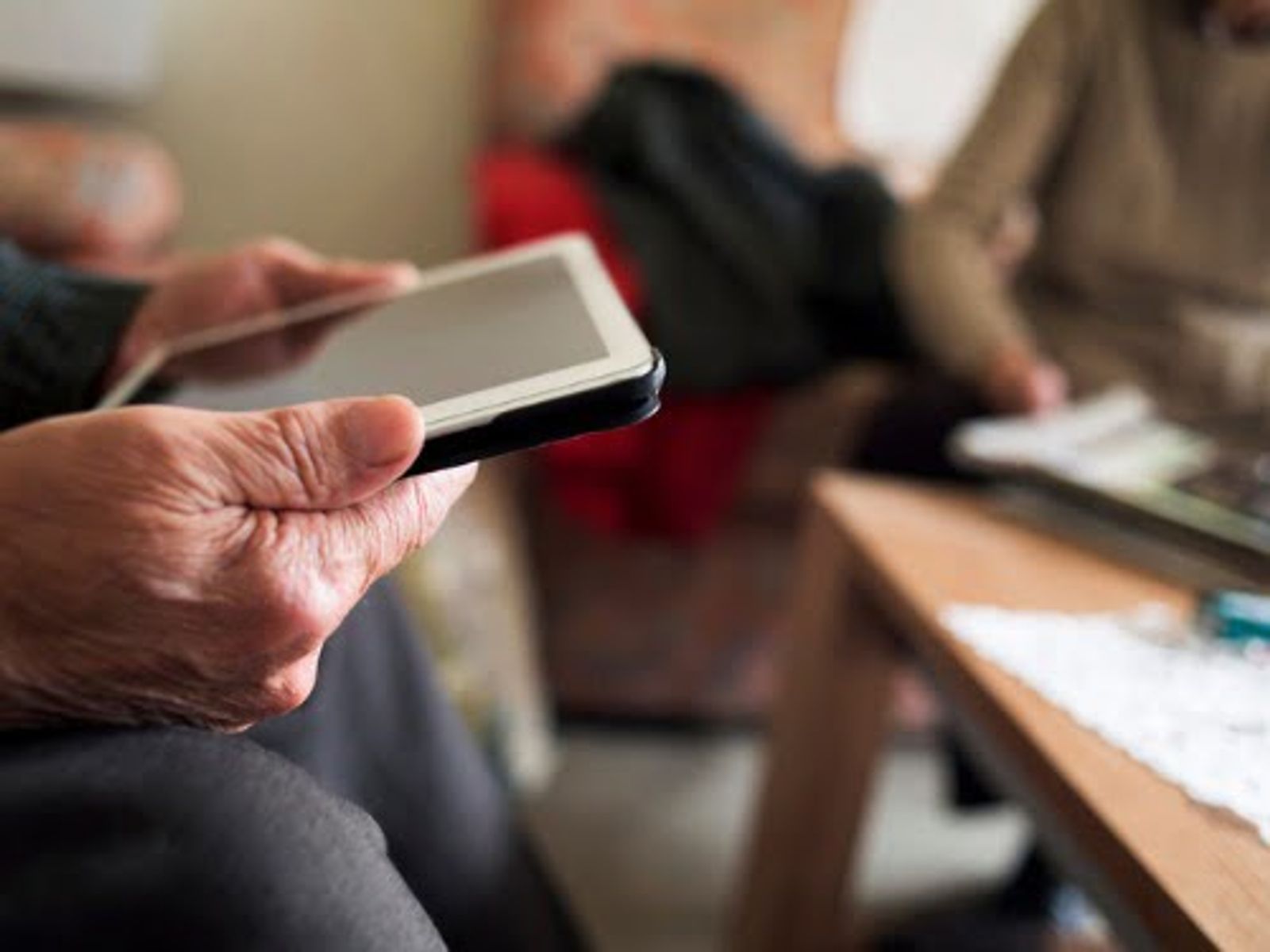 Person holding smartphone in foreground with blurred group of people in background at table.