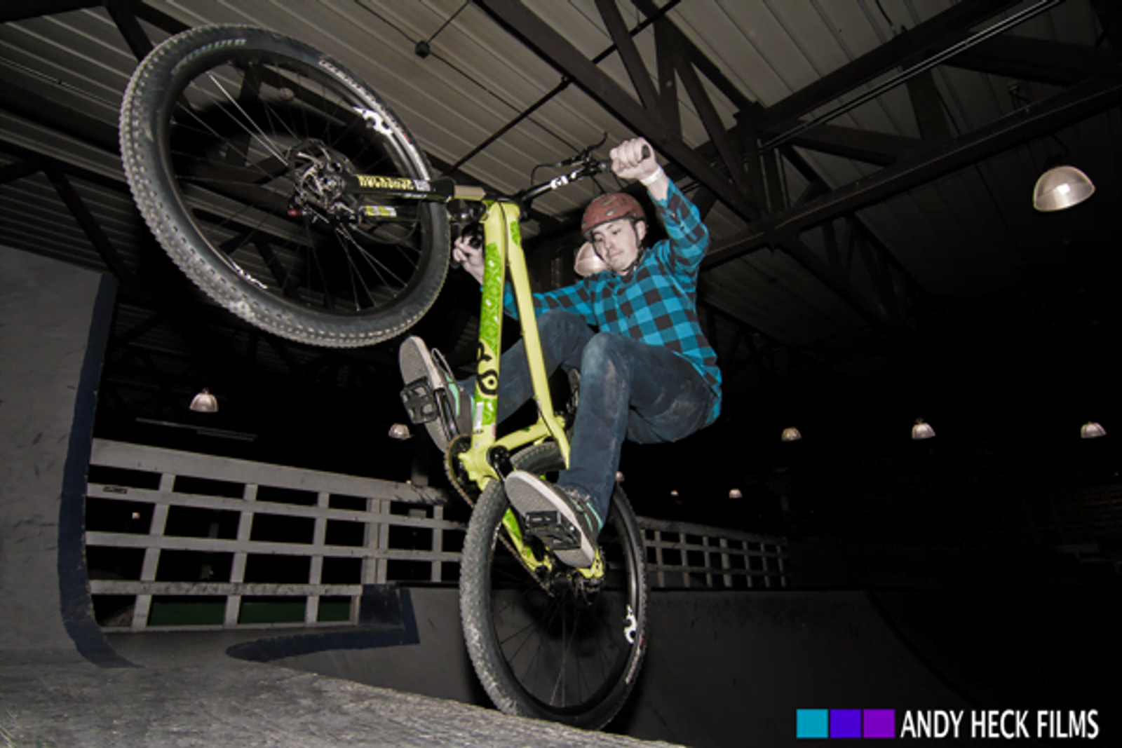 Cyclist performing wheelie with bike elevated indoors, wearing blue plaid shirt and helmet.