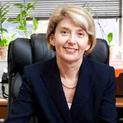 Mujer profesional de cabello rubio sentada en un escritorio de oficina, con un blazer oscuro y collar de perlas.