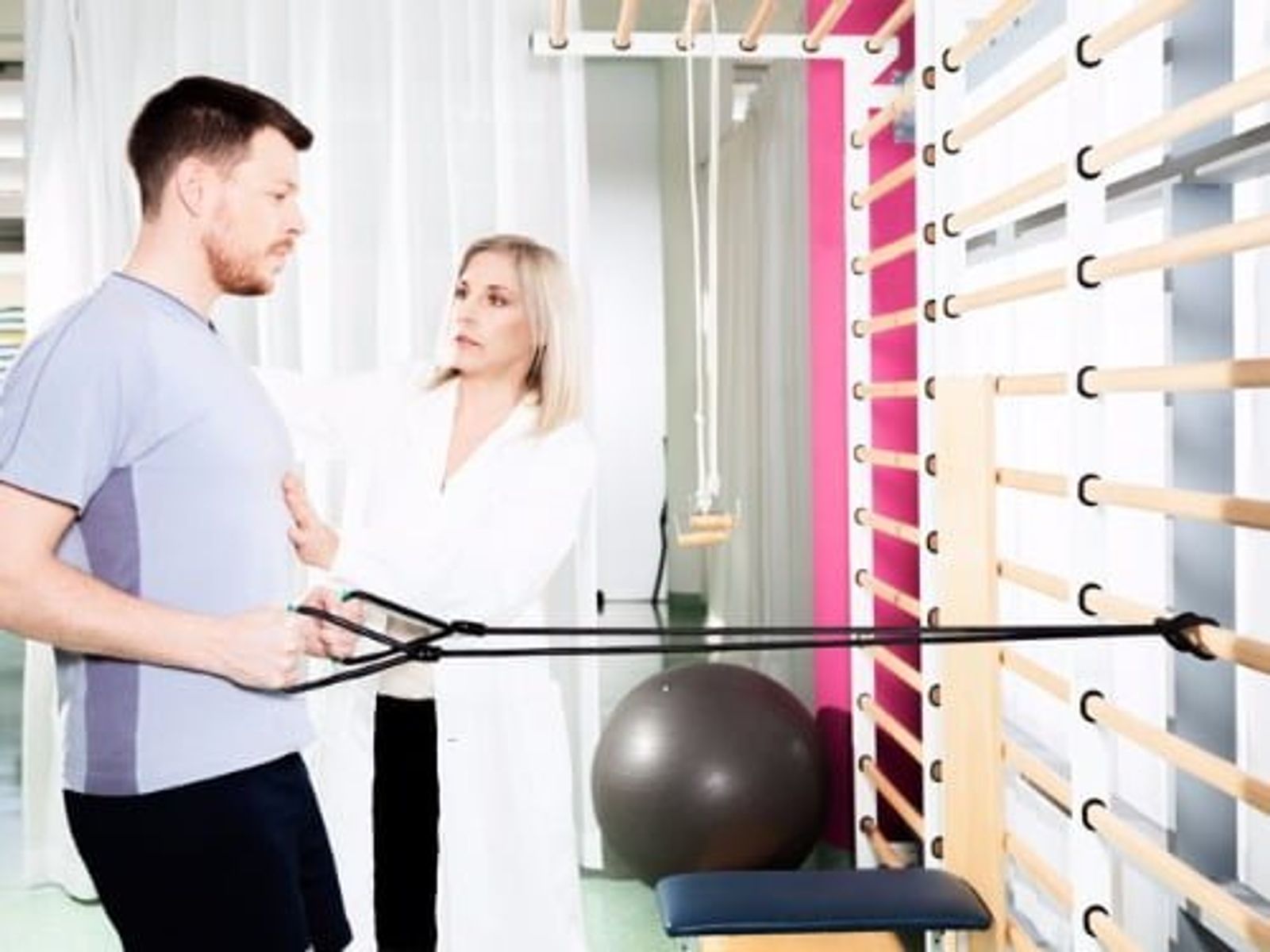 Physical therapist instructing male patient using resistance bands in clinic with exercise equipment.
