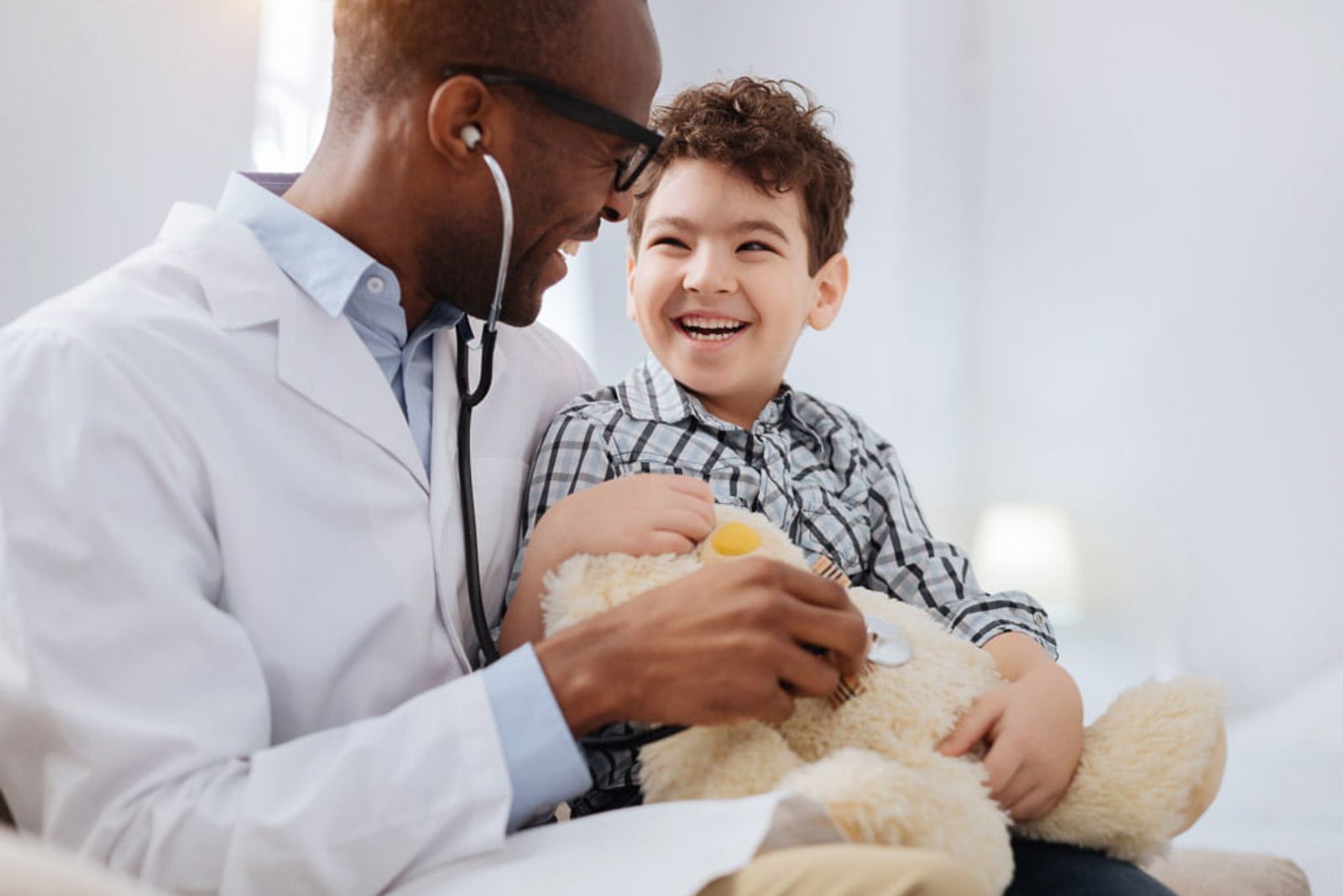 Médico examina a un niño sonriente con un estetoscopio y un oso de peluche.