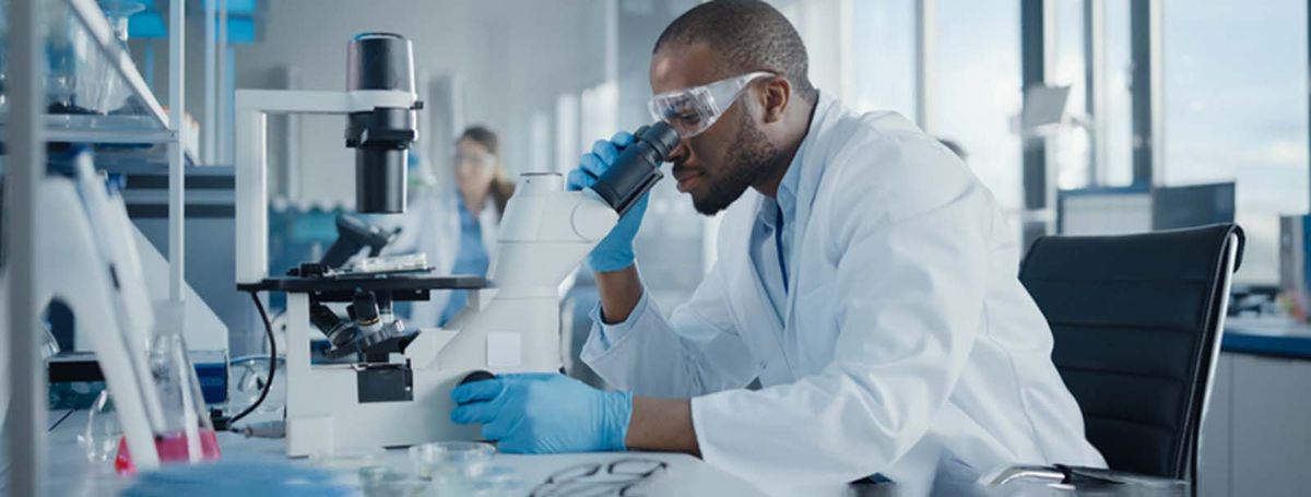 Científico con bata blanca y gafas de seguridad examina un portaobjetos de microscopio en un entorno de laboratorio moderno.
