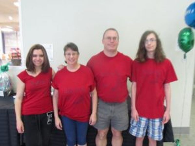 Four people wearing red shirts stand together indoors with balloons visible in background.