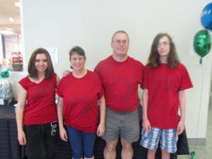 Four people wearing red shirts stand together indoors with balloons visible in background.