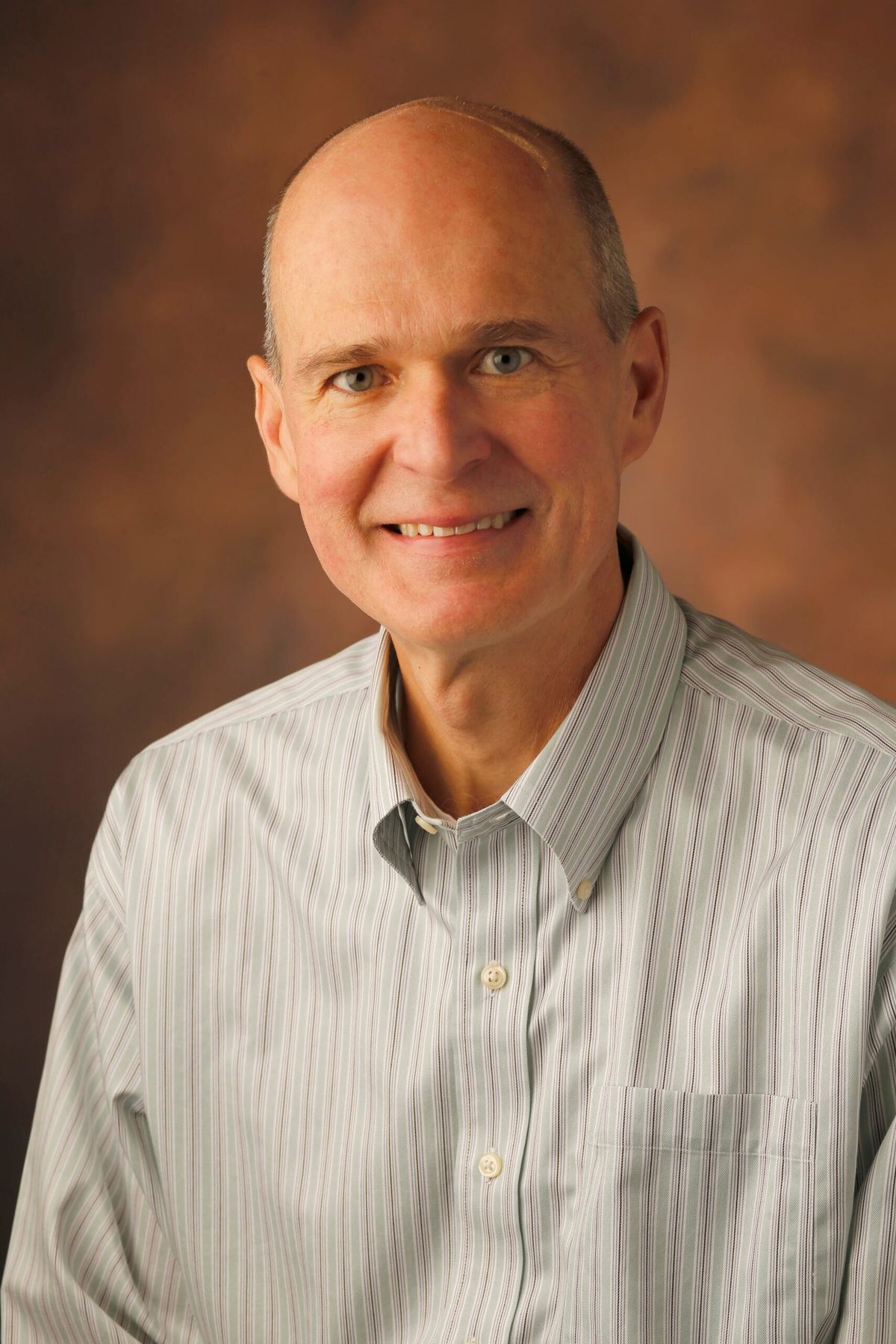Middle-aged man with receding hair wearing light gray striped button-up shirt against brown background.