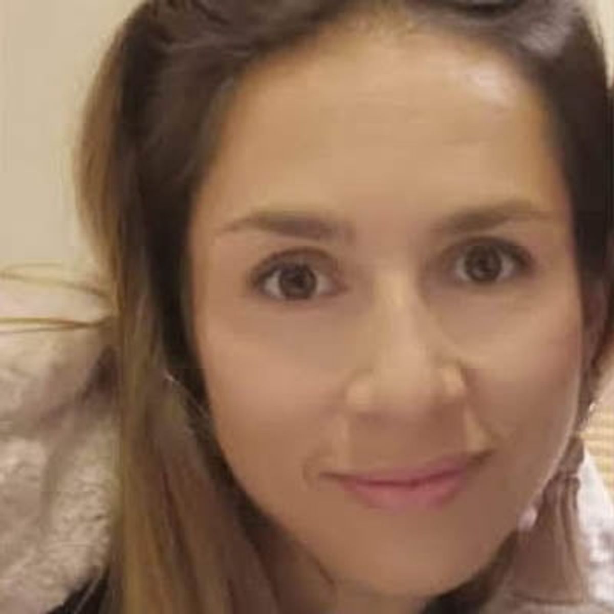 Close-up portrait of a woman with brown hair and pink lipstick, smiling at camera indoors.