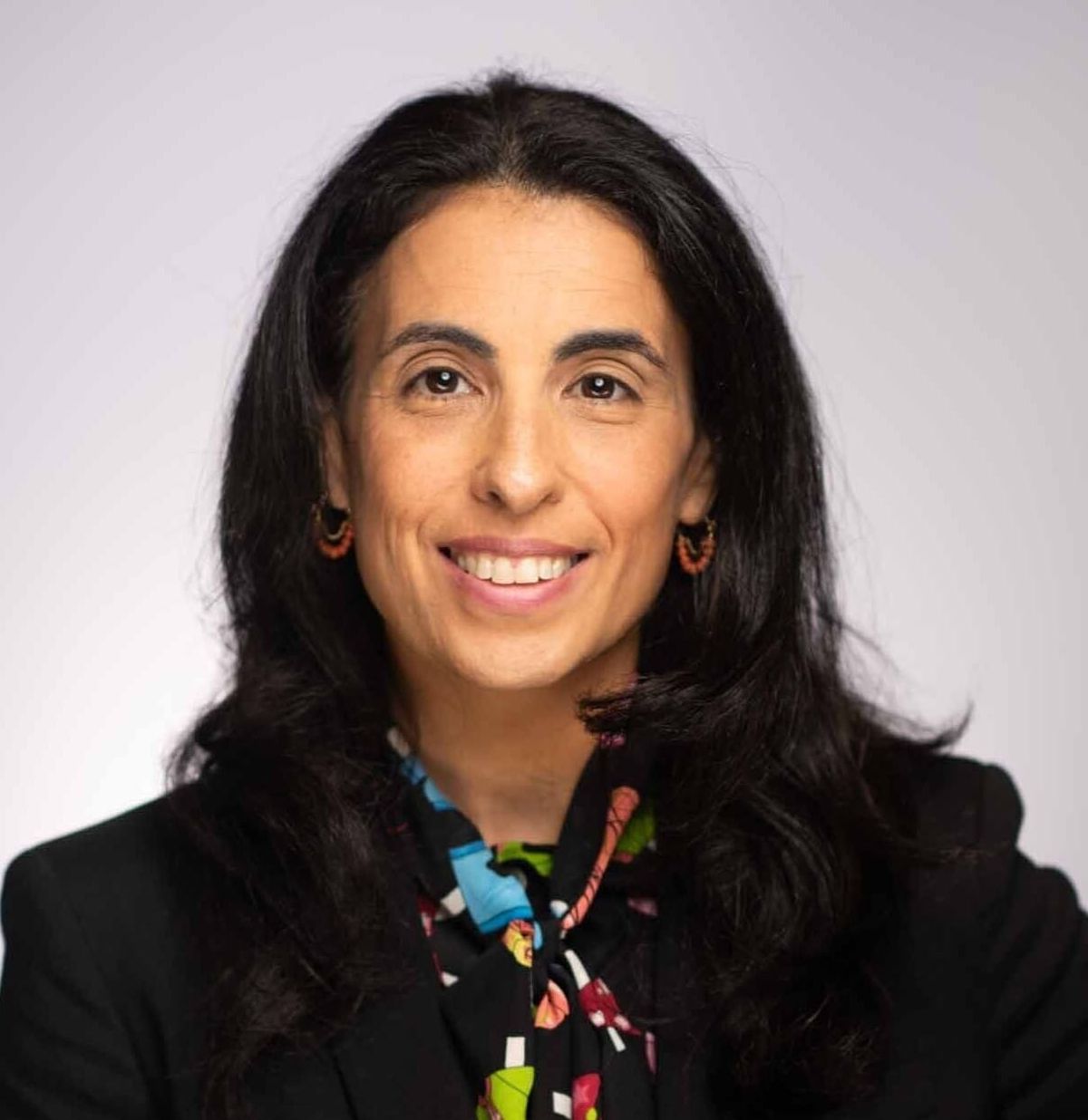 Professional headshot of a woman with long black hair, wearing a black blazer and colorful patterned scarf, smiling at the camera.
