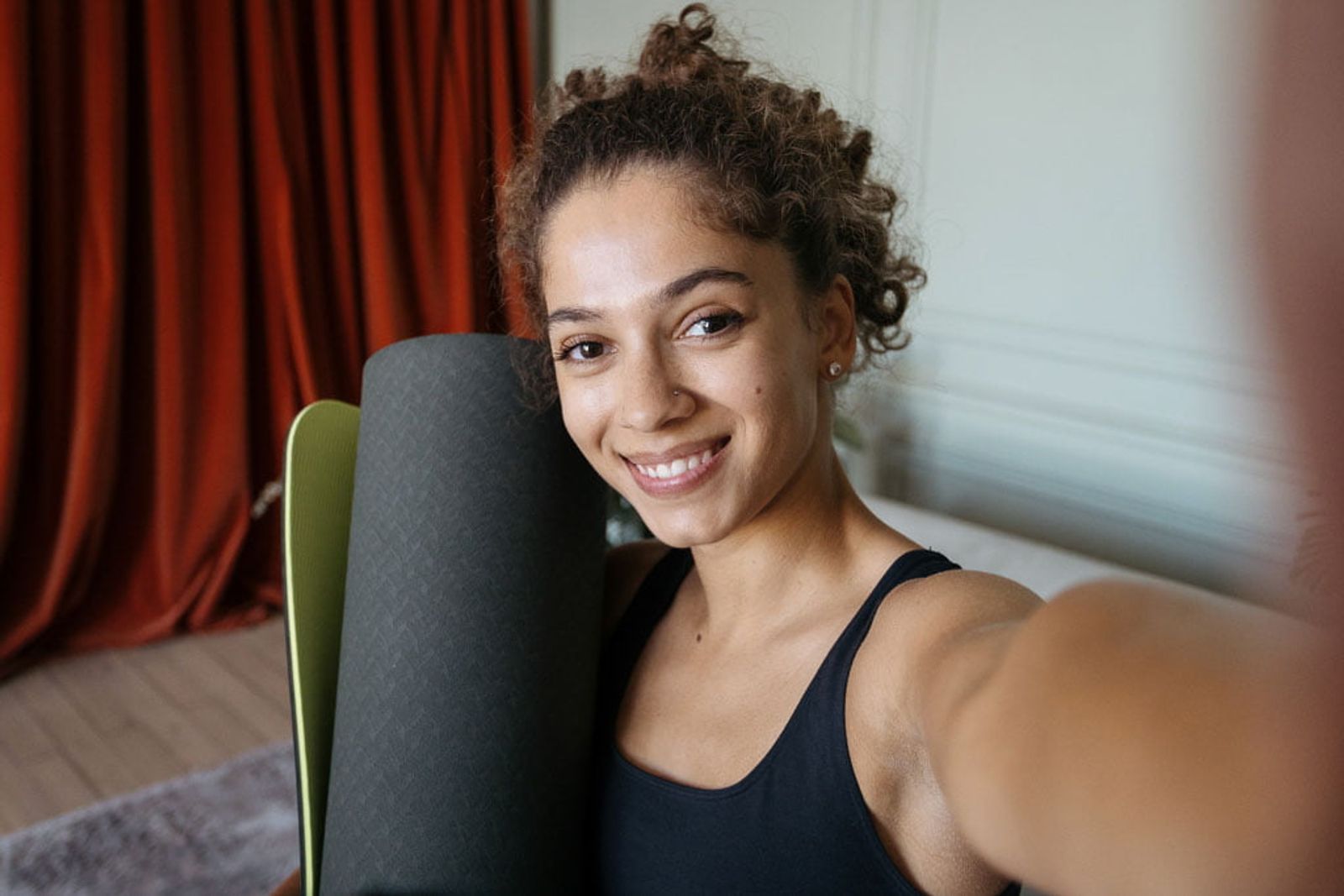 A smiling person in athletic wear holds a rolled-up yoga mat at home, ready to exercise.