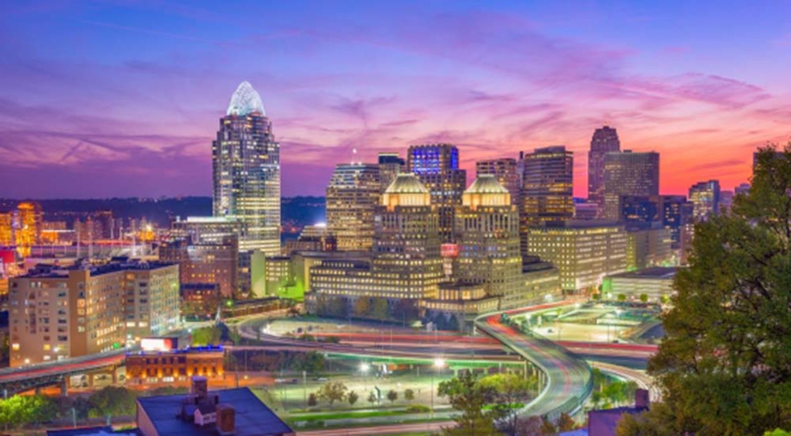 Horizonte de Cincinnati al atardecer, con edificios iluminados reflejados en el río.