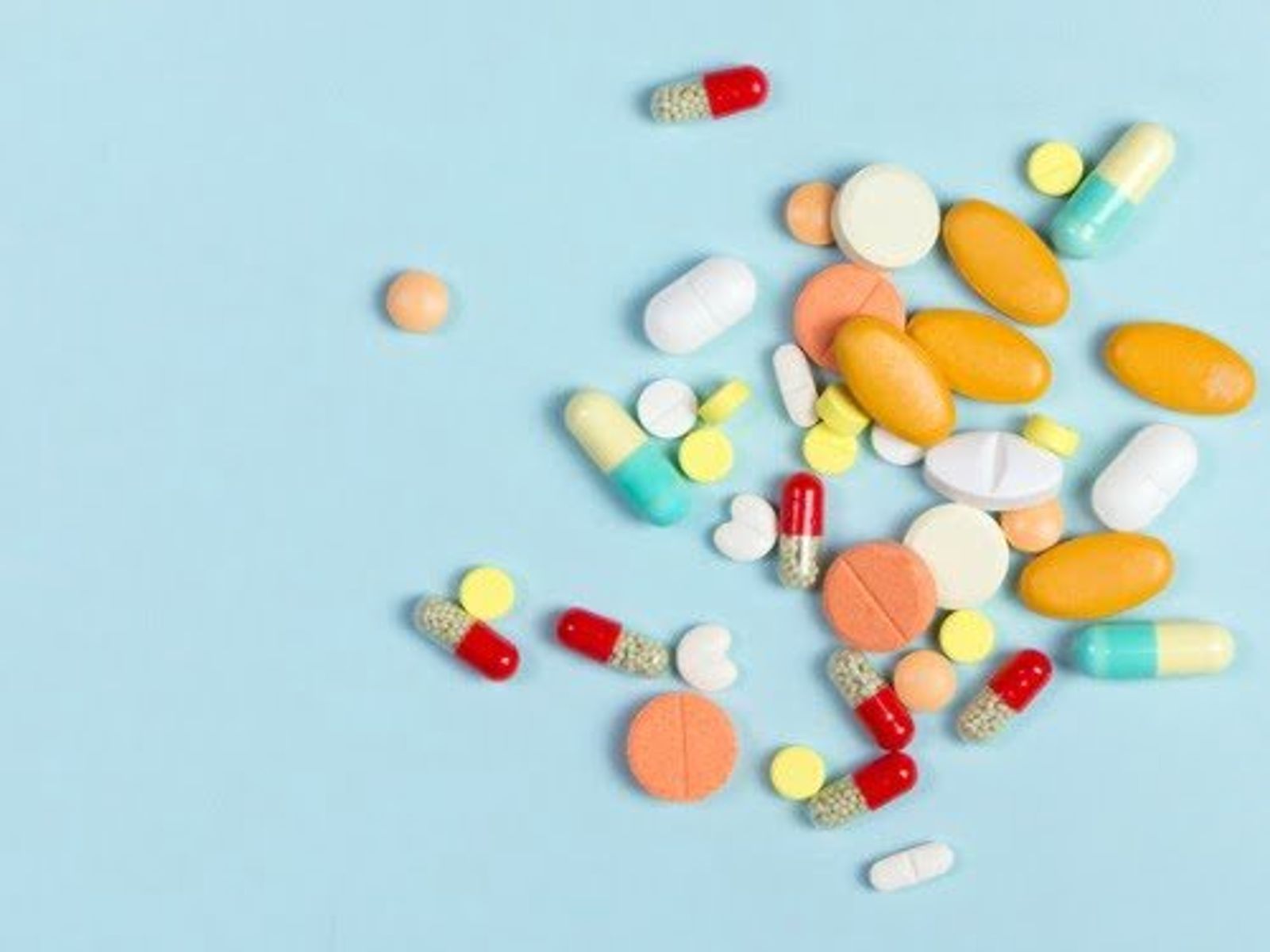 Assorted medications and pills scattered on light blue background, including capsules, tablets, and round pills in various colors.