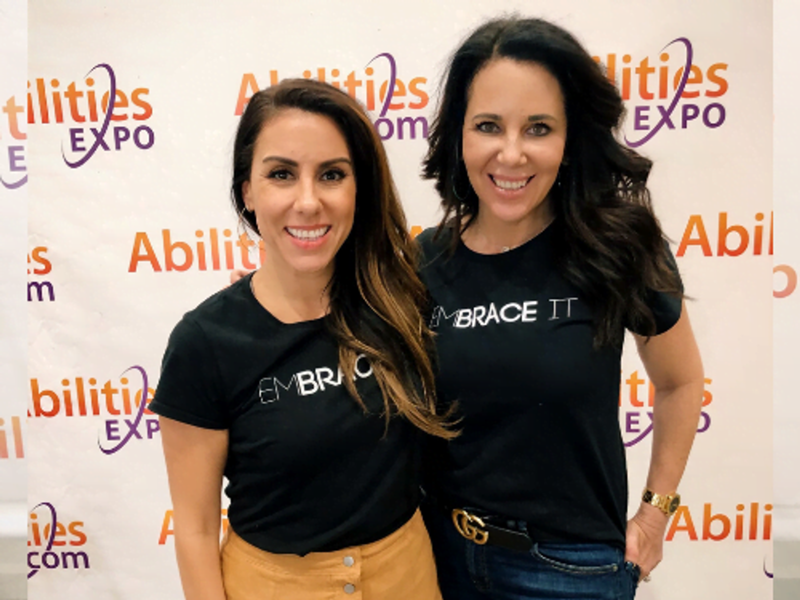 Dos mujeres con camisetas negras de EMBRACE están juntas frente a un fondo de Abilities Expo.