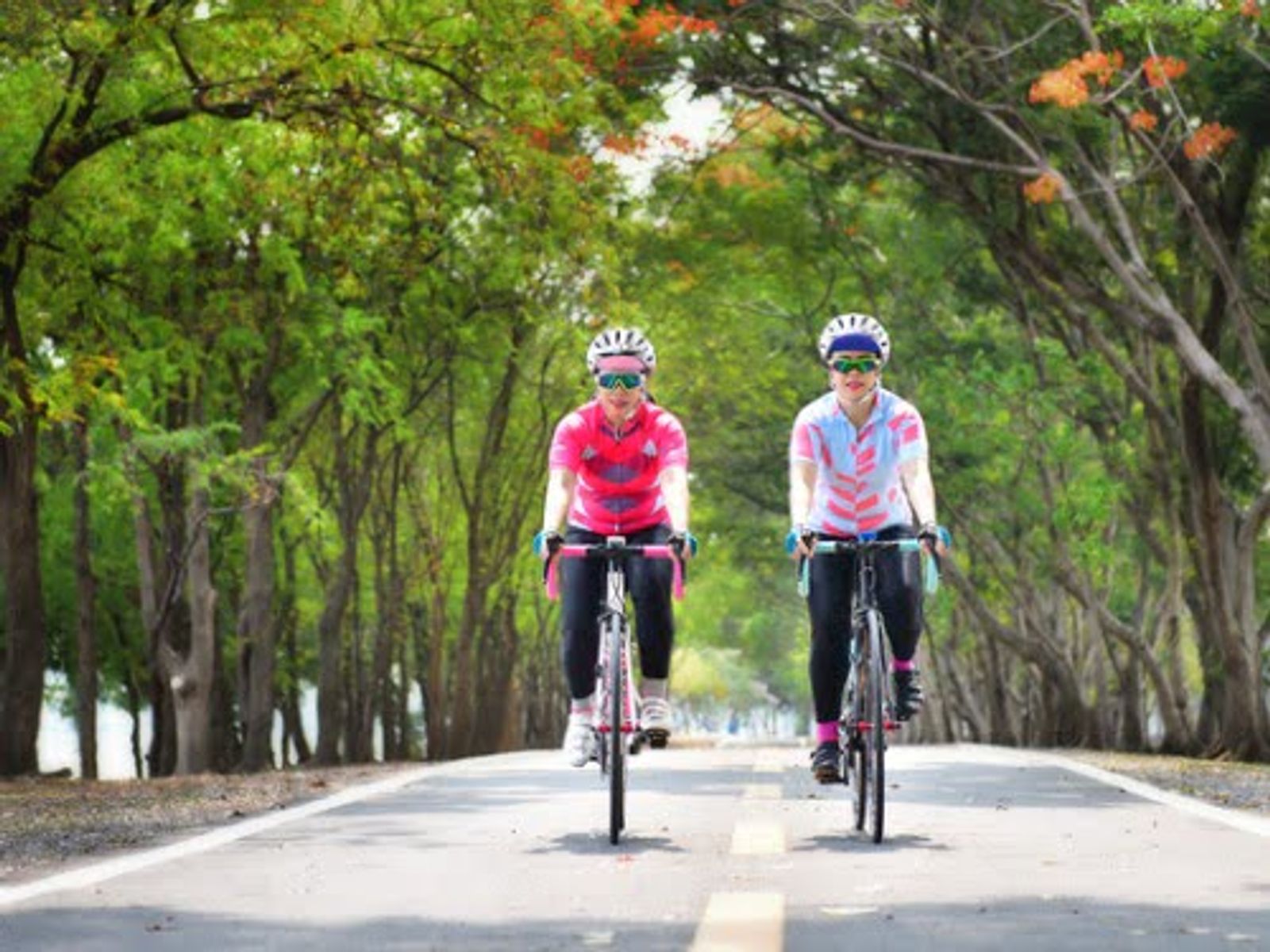 Two cyclists riding bikes on tree-lined road wearing helmets and athletic gear.