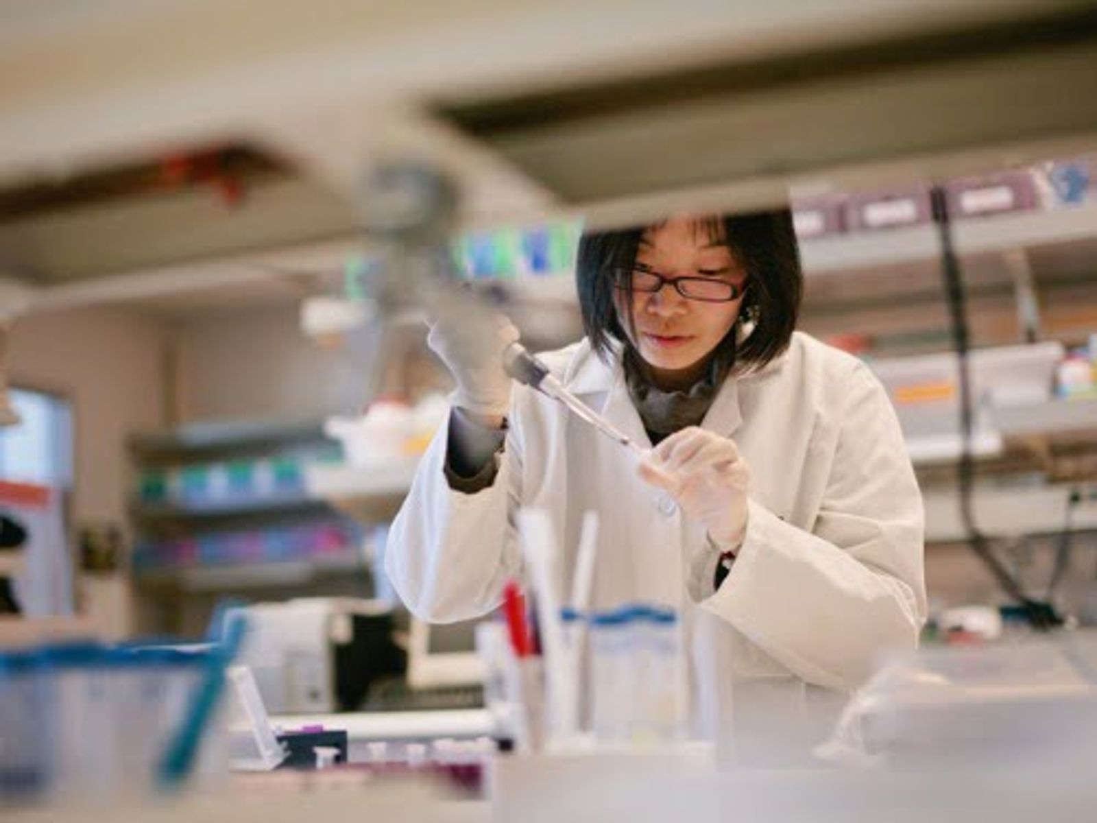 Scientist in white lab coat and glasses working with laboratory equipment at bench.