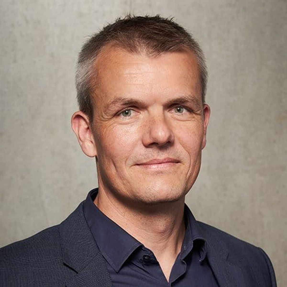 Professional headshot of a middle-aged man with short dark hair wearing a dark blue polo shirt against a neutral beige background.