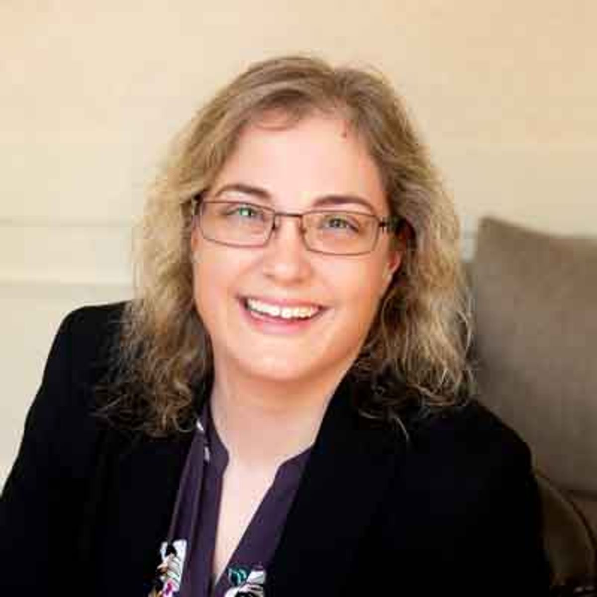 Woman with shoulder-length brown hair and glasses wearing a black blazer, smiling at camera indoors.